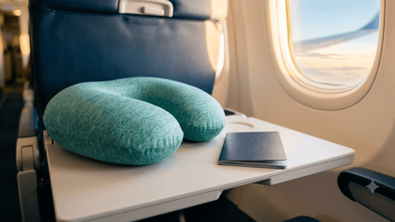A travel pillow and passport resting on an airplane tray table in warm window light.